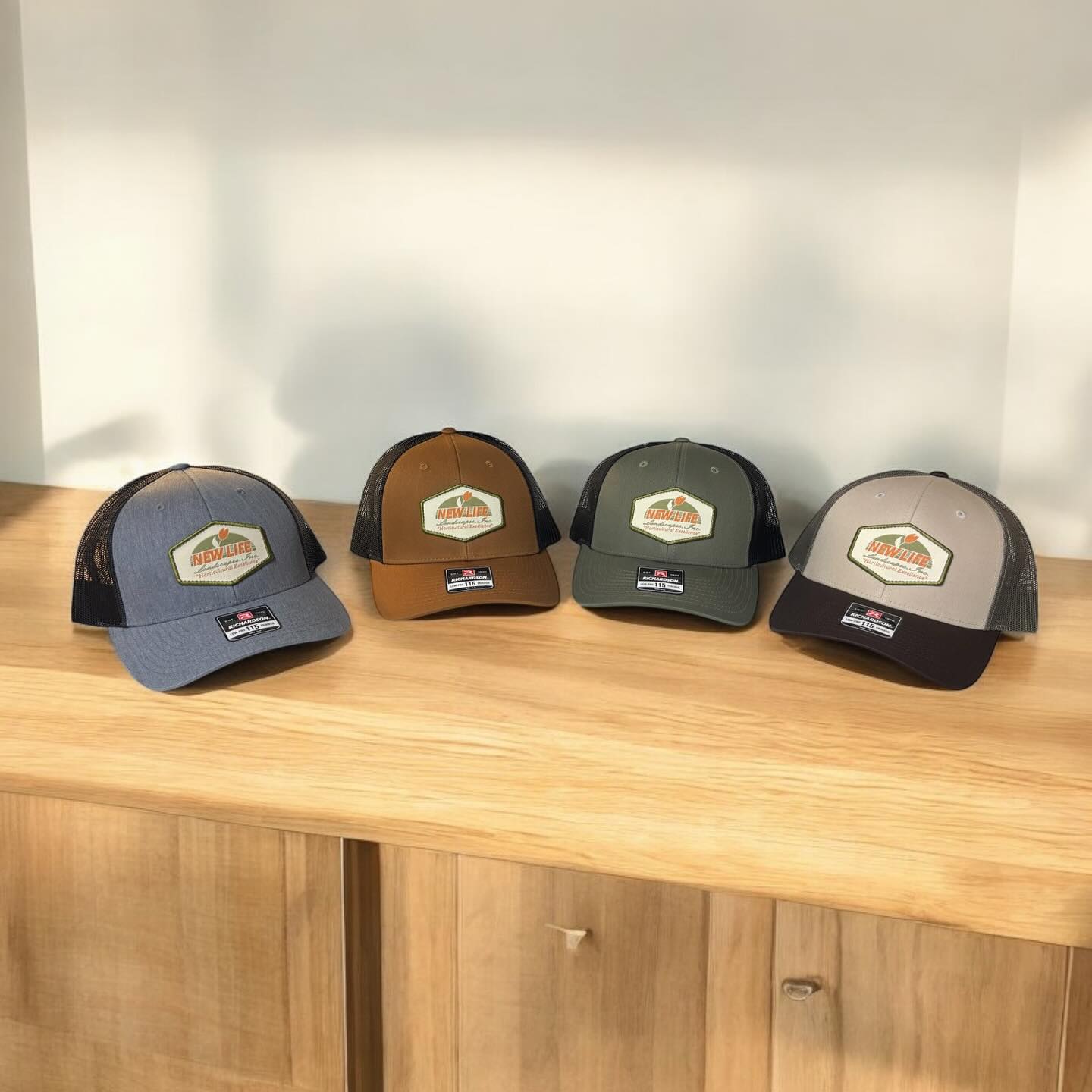 Four trucker hats in different colors with a leather patch featuring the New Life Landscaping logo.