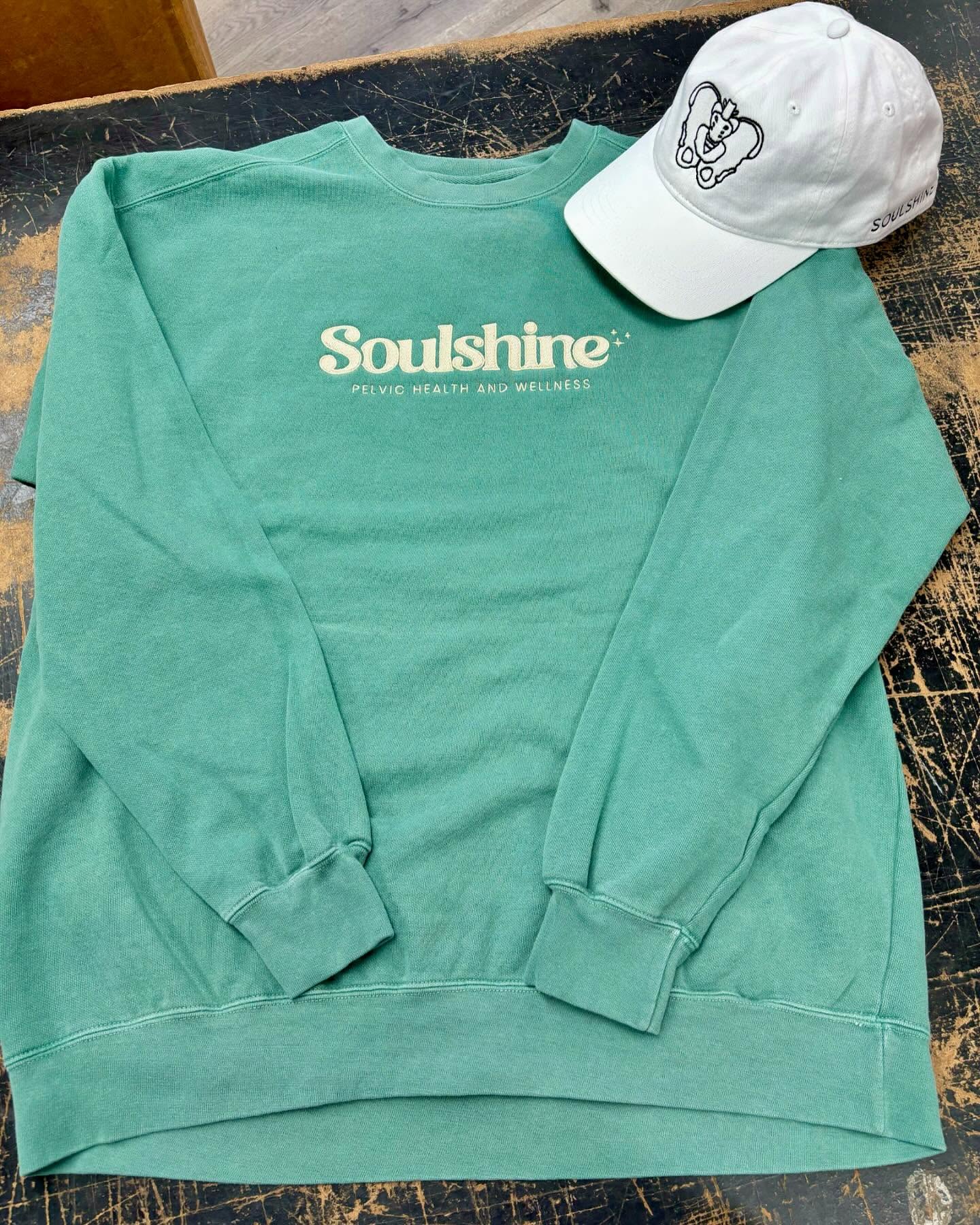 Green crewneck sweatshirt and white baseball cap featuring Soulshine Pelvic Health & Wellness branding.
