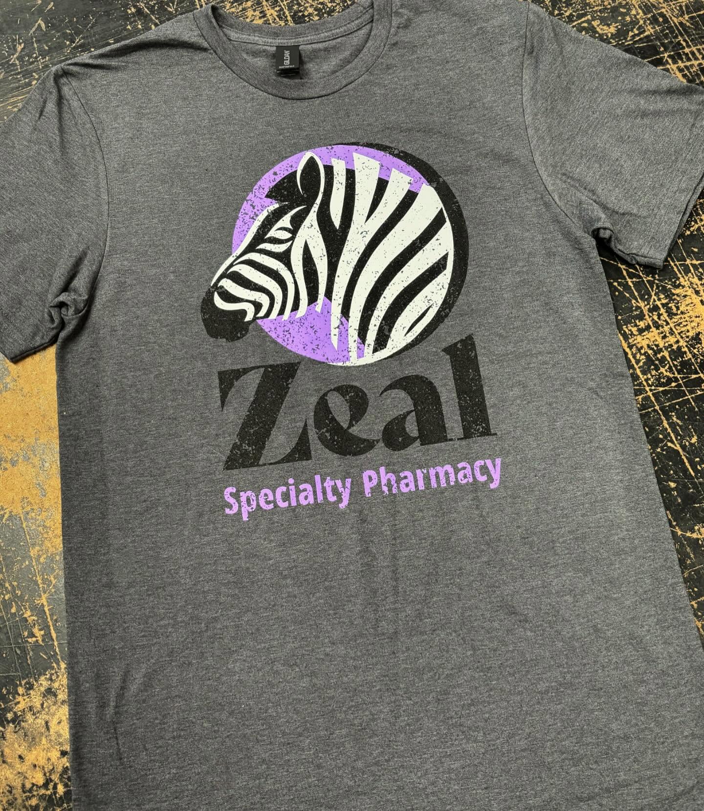 Gray T-shirt with a distressed zebra logo and purple Zeal Specialty Pharmacy branding.